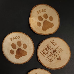 Puede incluir: Cuatro posavasos de madera con un diseño de huella de pata. Los posavasos están personalizados con los nombres "Bobi", "Paco", "Draco" y "Home is where my cats are".