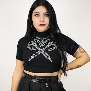 May include: A black cropped top with a white graphic of two crossed axes and stars. The top has a mock neck and short sleeves.