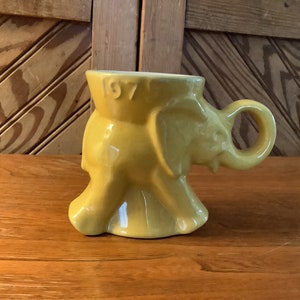 May include: Yellow ceramic elephant-shaped mug with a handle. The mug has the number "197" on the side. The mug is sitting on a wooden surface.