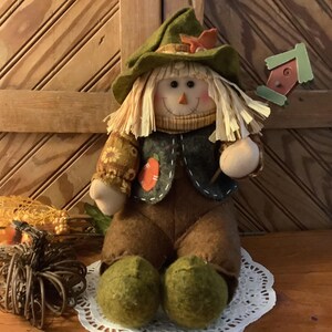 May include: A fabric scarecrow doll with straw hair, wearing a green hat, a dark vest with an orange pumpkin patch, and brown pants. The doll holds a small wooden birdhouse on a stick, and a decorative wire pumpkin sits nearby.