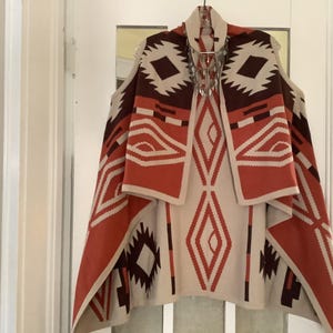May include: A sleeveless open-front cardigan featuring a geometric pattern in shades of cream, burnt orange, and dark brown. The knit garment has a draped silhouette with a wide collar and a repeating diamond and chevron motif throughout the fabric.