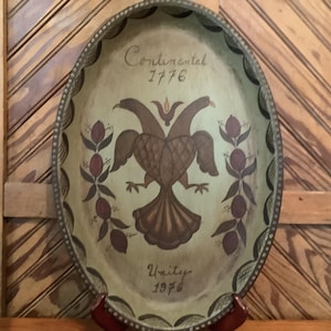 May include: An oval-shaped decorative tray with a light green background featuring a hand-painted brown double-headed eagle at the center. The text Continental 1776 is written above the eagle, and Unity 1976 is written below. Dark floral motifs flank the eagle.