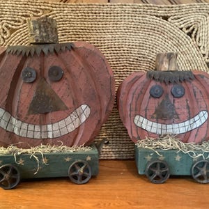 May include: Two carved wooden pumpkins with painted orange bodies, dark metal triangular noses, circular eyes, and wide white grins. Each pumpkin sits on a green wooden cart base with small metal wheels, decorated with painted stars and straw.