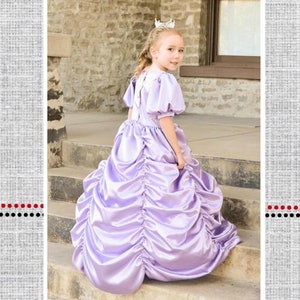 Könnte beinhalten: Ein junges Mädchen trägt ein lavendelfarbenes Kleid mit einem gerüschten Rock und einer Krone. Das Kleid heißt "Nancy's Fancy Ruched Dress" und ist Teil einer Kollektion namens "Polka Dot Umbrella".