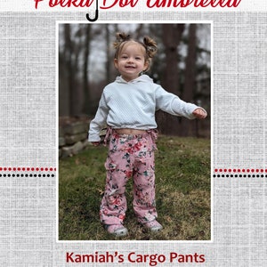 Puede incluir: Una niña pequeña con un suéter blanco y pantalones cargo de flores rosas. Los pantalones tienen una cintura con cordón y bolsillos a los lados. La niña está de pie en un área cubierta de hierba. El texto "Kamiah's Cargo Pants" está en la parte inferior de la imagen.