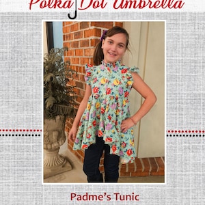 May include: A girl wearing a floral print tunic with a high neckline and ruffled sleeves. The tunic is a high-low style, with the front hemline shorter than the back. The girl is standing in front of a brick wall. The text "Padme's Tunic" is below the image.
