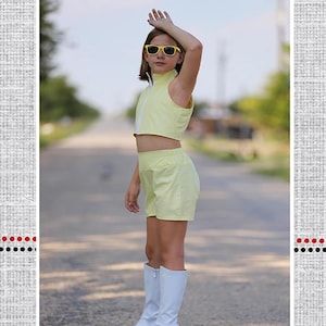 May include: A young person wearing a yellow crop top and shorts set, yellow sunglasses, and white boots. The image includes the text "Polka Dot Umbrella" and "Ramsey's Crop Top & Shorts Set". The background is a road.