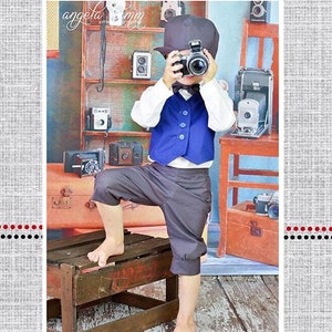 Könnte beinhalten: Ein kleiner Junge trägt eine blaue Weste, graue Hosen und einen schwarzen Hut und hält eine Kamera in der Hand. Er steht auf einer Holzkiste vor einem Regal mit Vintage-Kameras. Der Text "Tanner's Newsboy Knickers" befindet sich am unteren Rand des Bildes.