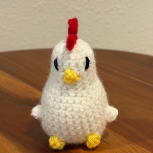 Pode incluir: Uma galinha de crochê branca feita à mão com uma crista vermelha, olhos pretos e bico e pés amarelos. O pequeno animal de pelúcia está sentado em uma superfície de madeira. A galinha tem pequenas asas e um corpo arredondado.