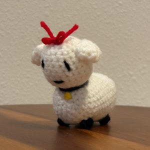 Pode incluir: Uma figura de cordeiro de crochê feita à mão. O cordeiro é branco com olhos, orelhas e pés pretos. Tem um laço vermelho na cabeça e uma coleira preta com uma etiqueta amarela. O cordeiro tem cerca de 8 cm de altura.