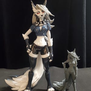 Puede incluir: Dos figuritas pintadas de zorros antropomorfos. La figurita más grande es blanca con detalles negros y ojos rojos. La figurita más pequeña es gris y tiene un diseño más detallado. Ambas figuritas están de pie con sus colas detrás de ellas.