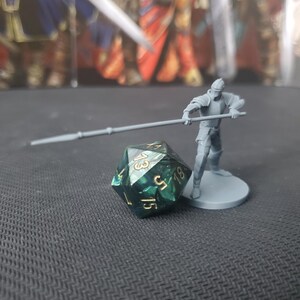 May include: A green and blue 20-sided die with gold numbers, showing a 15, next to a gray miniature figure holding a spear.