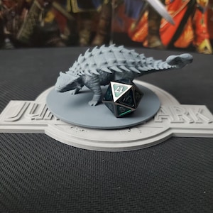 May include: A gray 3D-printed Ankylosaurus dinosaur model stands on a gray circular base, next to a multi-sided die. The base is set on a platform with the text "JURASSIC PARK". The background is blurred.