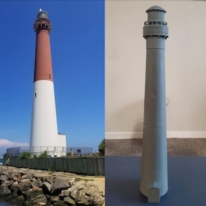 Puede incluir: Un faro alto con la parte superior roja y la base blanca contra un cielo azul claro. Junto a él, una maqueta gris del faro. El faro está en la costa, con rocas y una valla en primer plano.