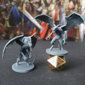 May include: Two gray painted miniature figurines of winged demons with horns. The larger demon is standing on a gray base with a gold and brown 20-sided die. The smaller demon is standing on a gray base.