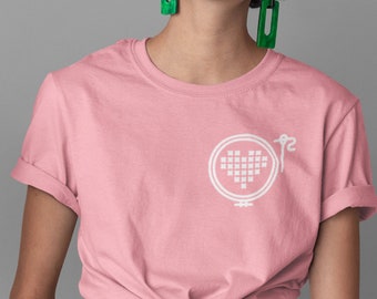 Op de afbeelding: Een roze t-shirt met een wit geborduurd ontwerp van een hart in een hoepel met een naald en draad.