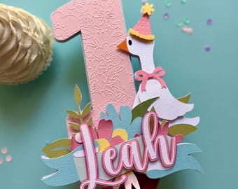 May include: A pink number one birthday cake topper with a floral design, a goose wearing a party hat, and the name "Leah" on a banner. The topper is decorated with flowers in shades of pink, blue, and green.
