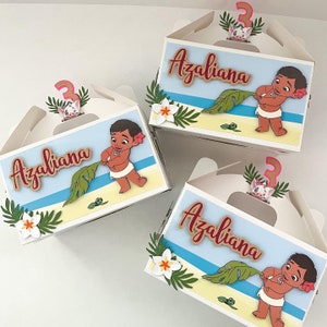 May include: Three white favor boxes with a blue and white beach scene. Each box has a brown paper cutout of the name "Azaliana" and a pink number "3". The boxes feature a cartoon image of a brown-skinned baby holding a green leaf.