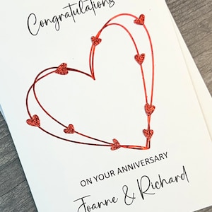 May include: A white card with the text "Congratulations" in black cursive writing. A red glitter heart is drawn on the card with smaller red glitter hearts along the lines. The text "On your anniversary Joanne & Richard" is written in black cursive writing below the heart.