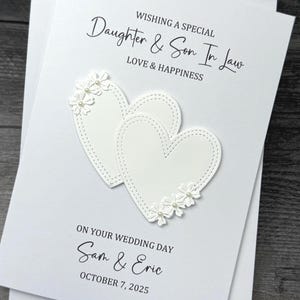 May include: A white wedding card with the text "WISHING A SPECIAL Daughter & Son In Law LOVE & HAPPINESS ON YOUR WEDDING DAY Sam & Eric OCTOBER 7, 2025". Two white hearts with floral embellishments are featured.