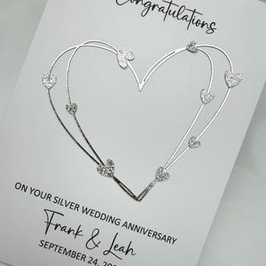 May include: A white greeting card with the word "Congratulations" in black script. A silver heart design with sparkling heart accents is featured. The card reads "ON YOUR SILVER WEDDING ANNIVERSARY" and includes the names "Frank & Leah" and the date "SEPTEMBER 24, 2000."