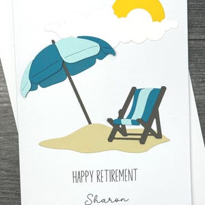 May include: A white card with a blue and white beach chair, a blue and white umbrella, a white cloud, and a yellow sun. The text "HAPPY RETIREMENT Sharon" is printed on the card.