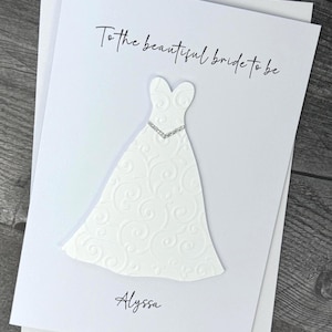 May include: A white card with the text "To the beautiful bride to be" and the name "Alyssa". The card features a white, textured, die-cut wedding dress with a rhinestone-like embellishment.