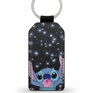 May include: A blue Stitch character from Lilo & Stitch is holding a pink bubble with a starry night background. The keychain is made of black faux leather with a silver key ring.