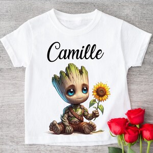 Peut inclure: T-shirt blanc avec un personnage de dessin animé tenant un tournesol, avec le nom "Camille" en écriture noire. Le personnage a une apparence en bois avec des yeux bleus. Des roses rouges sont au premier plan.