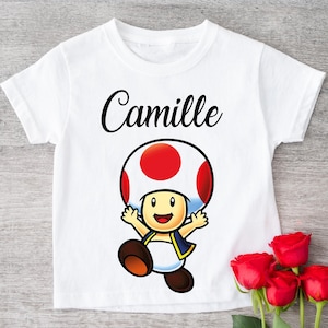 Könnte beinhalten: Weißes T-Shirt mit einer Cartoonfigur mit rotem und weißem Pilzhut, Arme ausgestreckt. Der Name "Camille" steht in Schreibschrift über der Figur. Rote Rosen in der unteren rechten Ecke.