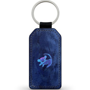 May include: A blue leather keyring with a silver key ring and a blue and pink lion silhouette graphic. The keyring has a distressed look and the graphic is slightly faded.