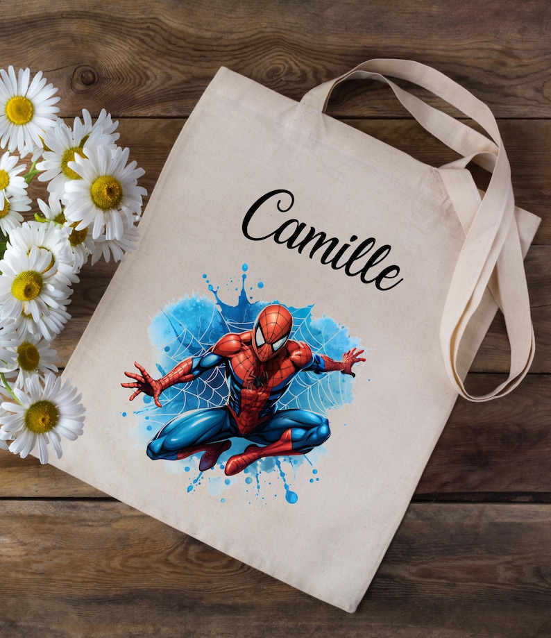 May include: A white canvas tote bag with a blue and red Spiderman graphic and the name "Camille" printed in black cursive.