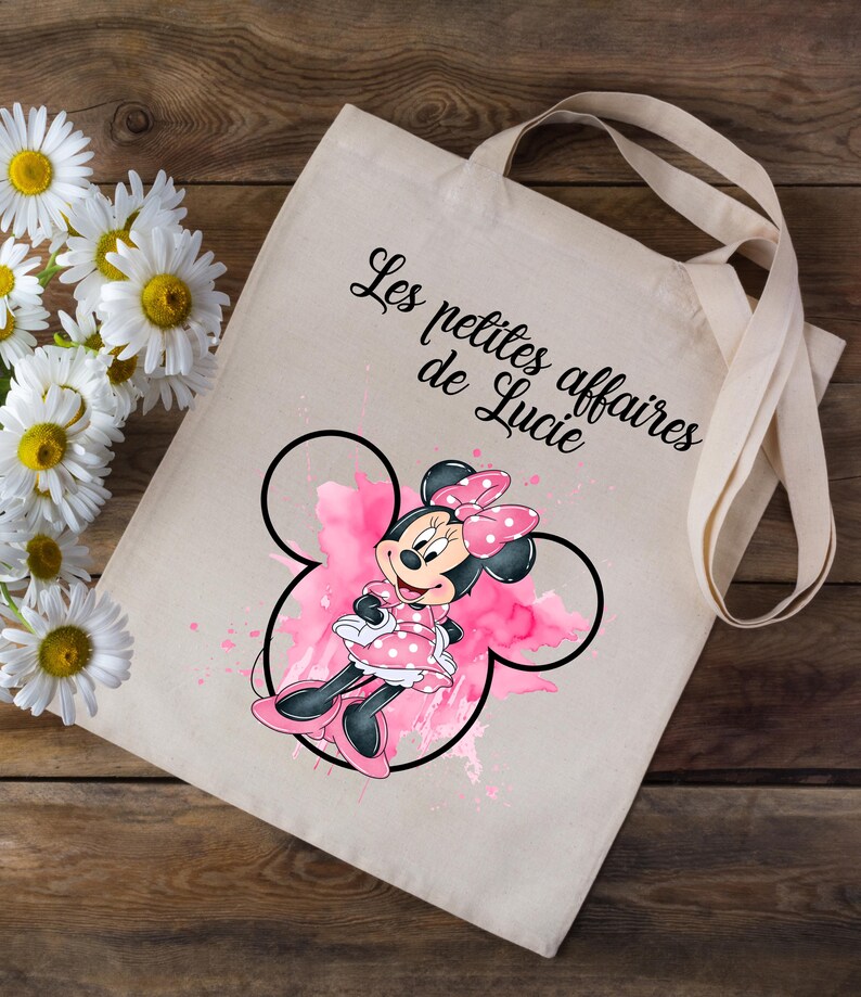 Może przedstawiać: Biała torba typu tote z akwarelową ilustracją Minnie Mouse w kolorze r&oacute;żowym i czarnym. Na torbie nadrukowany jest tekst "Les petites affaires de Lucie".