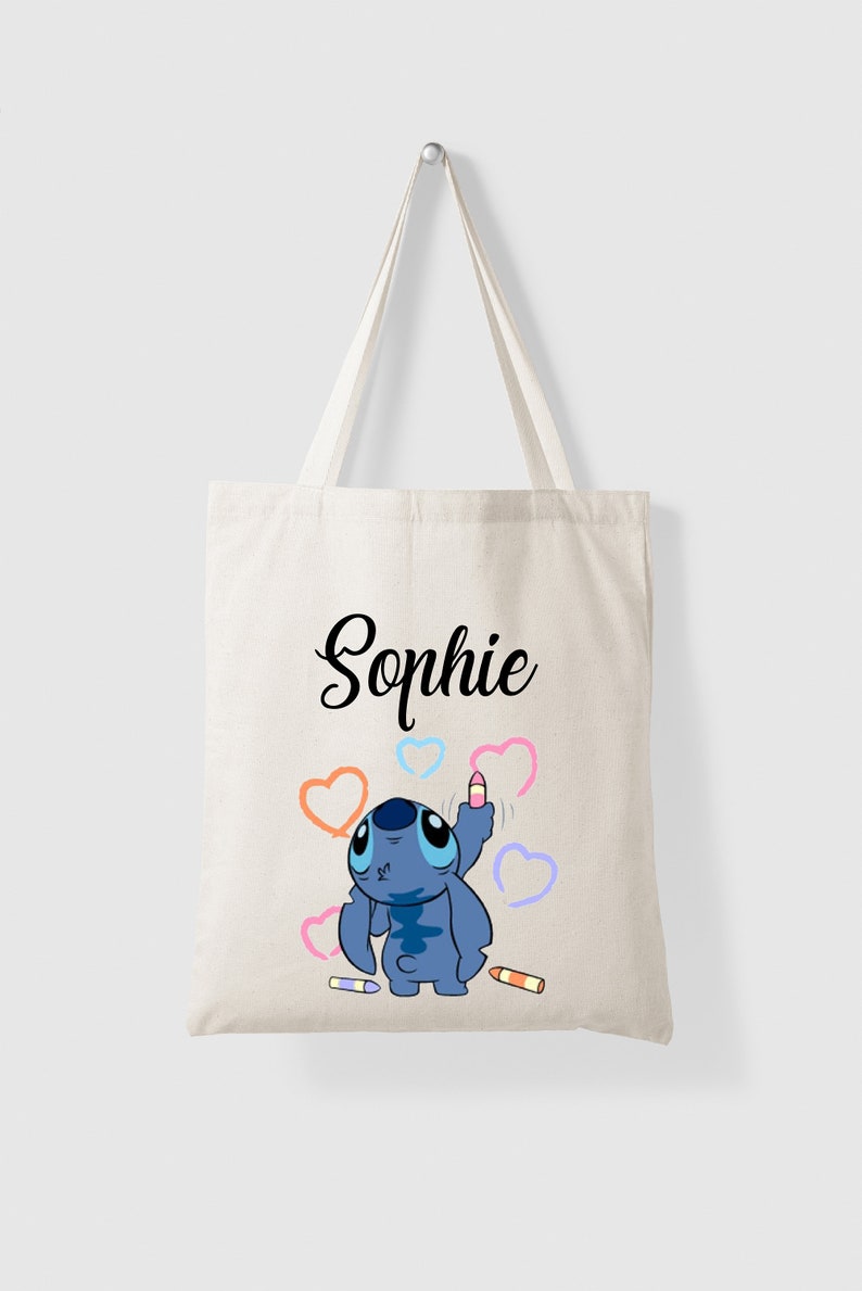 Puede incluir: Una bolsa de lona blanca con un personaje azul Stitch dibujando corazones con un cray&oacute;n rosa. La bolsa tiene el nombre "Sophie" impreso en cursiva negra.