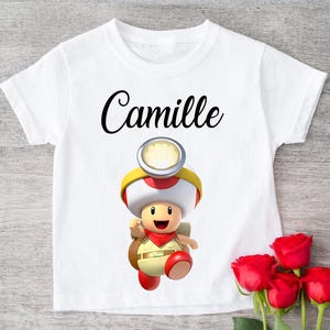 Peut inclure: T-shirt blanc avec un personnage de dessin animé portant un chapeau champignon rouge et blanc, un gilet rouge et un sac à dos marron. Le nom "Camille" est écrit en script noir au-dessus du personnage. Des roses rouges sont dans le coin inférieur droit.