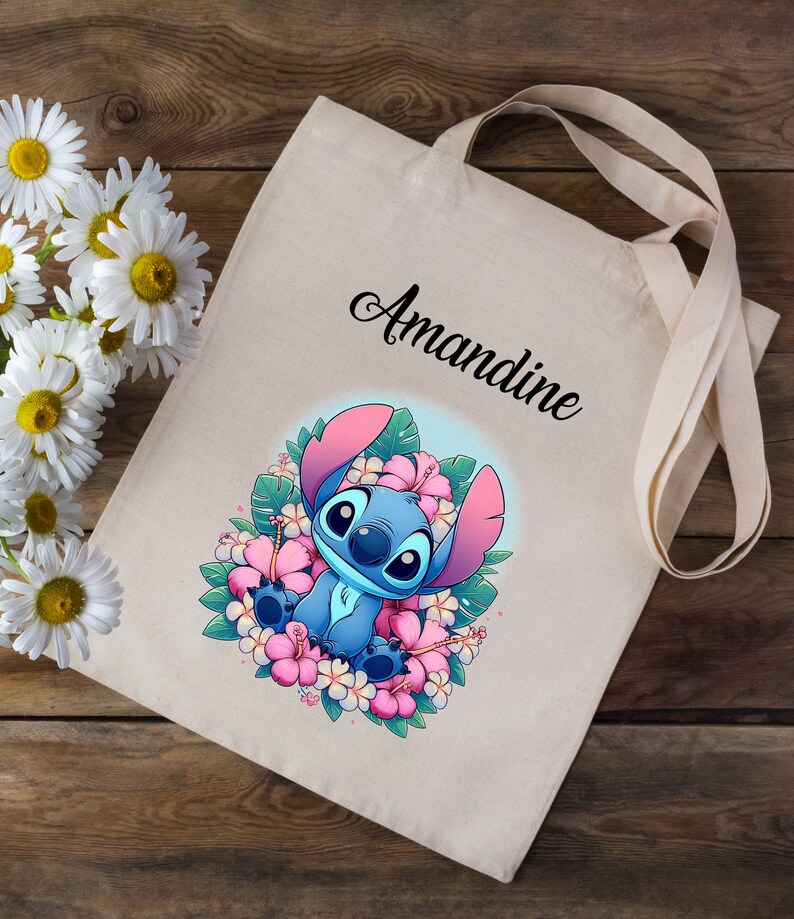 May include: A beige tote bag with a blue Stitch character from Lilo & Stitch surrounded by pink flowers. The bag has the name "Amandine" printed on it.