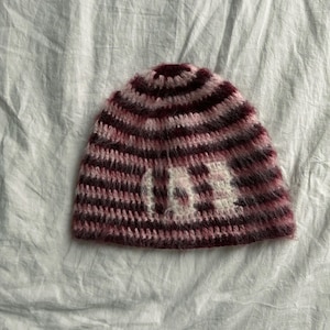 Op de afbeelding: Een handgemaakte, gestreepte beanie in tinten bordeaux, roze en wit. De hoed heeft het woord "I AM" in witte wol over de voorkant. De beanie is gemaakt van een zacht, getextureerd garen.