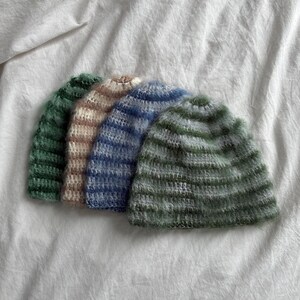 Op de afbeelding: Vier gebreide mutsen in groen, beige, blauw en grijs zijn in een rij gerangschikt. De hoeden zijn gemaakt van zacht, getextureerd garen en worden gepresenteerd op een witte, gekreukelde stof. De mutsen hebben een horizontaal gestreept patroon.