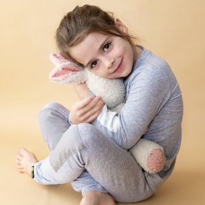 Puede incluir: Una niña pequeña con pijama gris está sentada sobre un fondo beige. Está abrazando un conejito de peluche blanco con orejas rosas cerca de su rostro. Está sonriendo a la cámara.