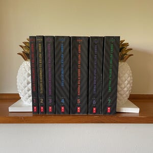 May include: A complete seven-volume set of Harry Potter books with black spines and colorful text, arranged between two white and gold pineapple-shaped bookends. Each spine displays the title, author name, and a large number from one to seven.