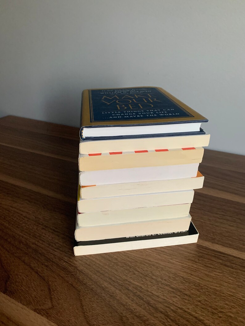 Pu&ograve; includere: Una pila di libri su una superficie di legno. Il libro in cima ha una copertina dorata con il titolo "Make Your Bed" e il testo "Little things that can change your life...and maybe the world."