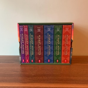 May include: A seven-volume Harry Potter book set housed in a rectangular cardboard slipcase. The spines are colored purple, magenta, teal, maroon, blue, green, and orange, each featuring the title, author name, and a numbered year icon.