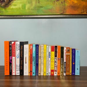 May include: A bookshelf filled with a variety of books, including titles like "Sapiens", "Think Again", "Shoe Dog", "The Magic of Thinking Big", "Mindset", "Start With Why", "Leaders Eat Last", and "The 48 Laws of Power".