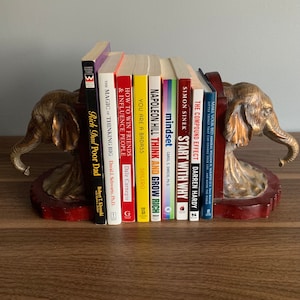 Pu&ograve; includere: Un paio di ferma libri a forma di elefante dorati che tengono una collezione di libri di auto-aiuto con titoli come "Pensa e arricchisci te stesso", "Inizia con il perch&eacute;", "Mentalit&agrave;" e "L'effetto composto".