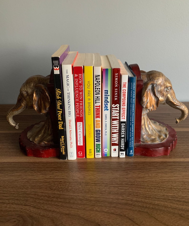 Pu&ograve; includere: Un paio di ferma libri a forma di elefante dorati che tengono una collezione di libri di auto-aiuto con titoli come "Pensa e arricchisci te stesso", "Inizia con il perch&eacute;", "Mentalit&agrave;" e "L'effetto composto".
