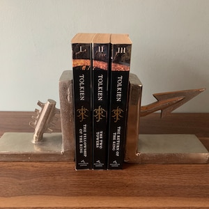 May include: Three books from the Lord of the Rings trilogy, held by a silver-colored bookend. The bookend has a key design on one side and an arrow on the other. The books are titled "The Fellowship of the Ring", "The Two Towers", and "The Return of the King".
