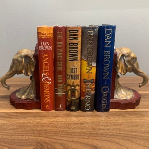 Op de afbeelding: Een set boekenleggers met olifantensculpturen met een rode houten basis. De boekenleggers houden een verzameling van vijf boeken van Dan Brown vast, waaronder "De Da Vinci Code", "Het Verloren Symbool", "Inferno" en "Origin".