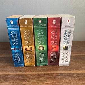 May include: A collection of five books by George R.R. Martin, including "Game of Thrones" and "A Dance with Dragons." The book spines are various colors, with titles and author's name in white. The books are displayed on a wooden surface.