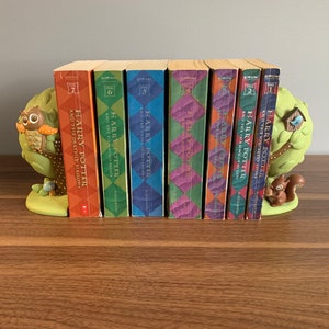 May include: A set of seven Harry Potter books, with colorful covers, are displayed on a wooden shelf with owl and squirrel bookends. The books are arranged in order from the first book, "Harry Potter and the Sorcerer's Stone", to the seventh book, "Harry Potter and the Deathly Hallows".
