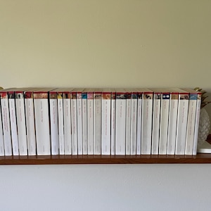 May include: A row of twenty-six white-spined Oxford World's Classics books arranged vertically on a wooden shelf. The books are held in place by two white pineapple-shaped bookends with gold-colored tops, positioned at each end of the collection.